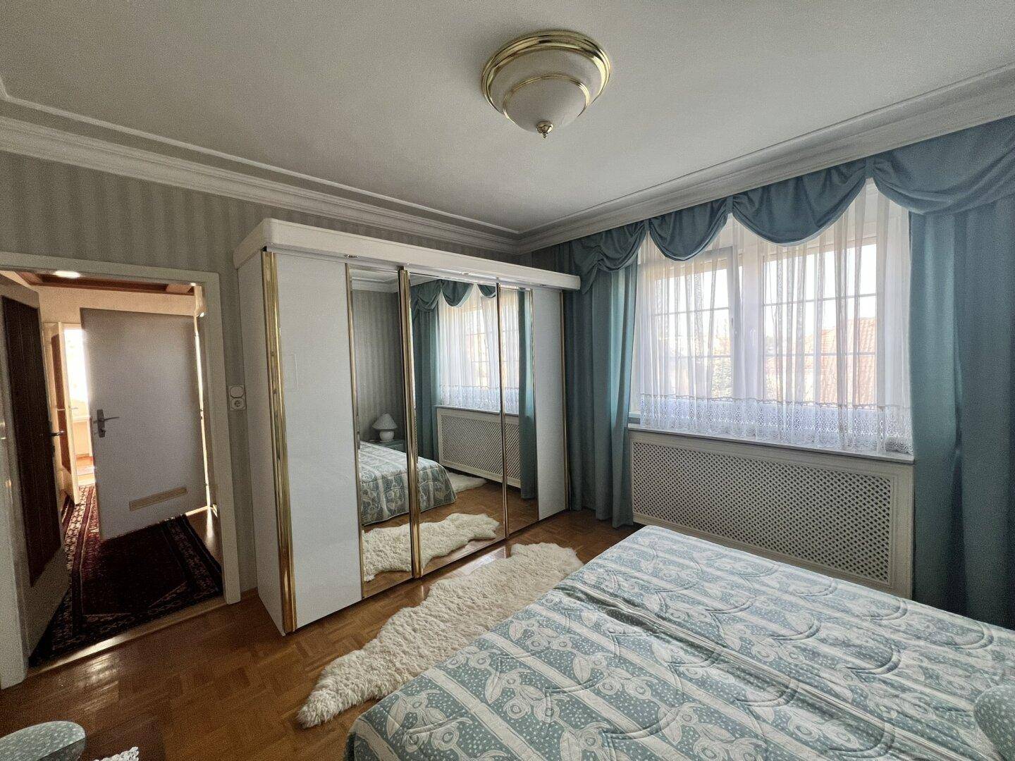 Schlafzimmer mit Spiegelschrank, Holzboden und hellen Fenstern mit blauen Vorhängen.