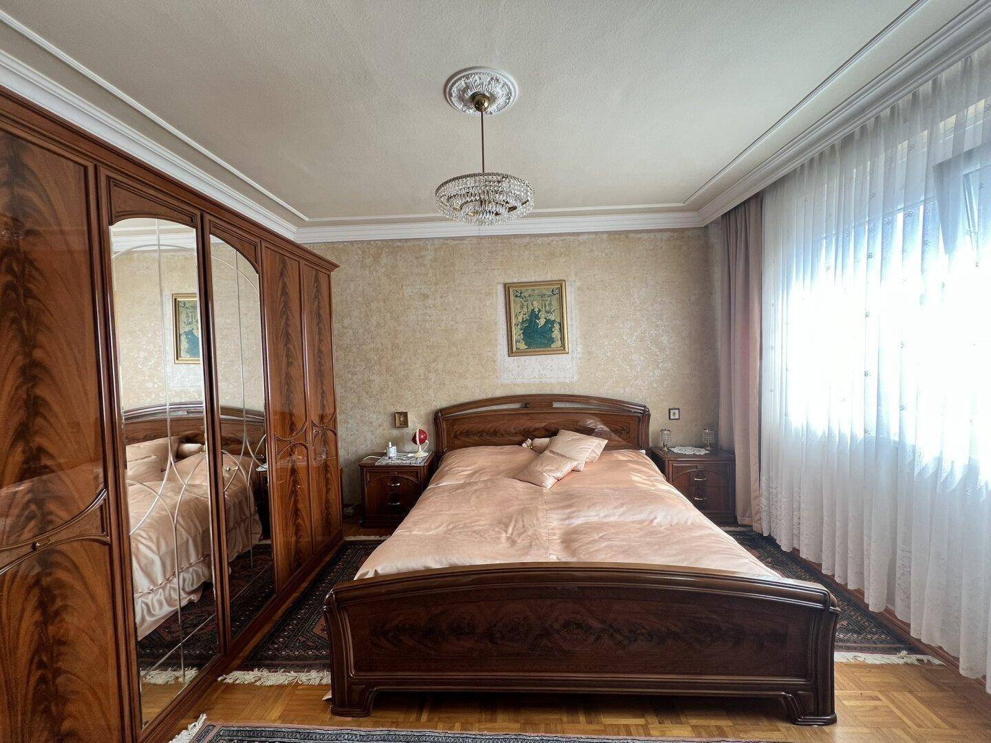 Helles Schlafzimmer mit Holzbett, Spiegelschrank und Fenster mit hellen Vorhängen.