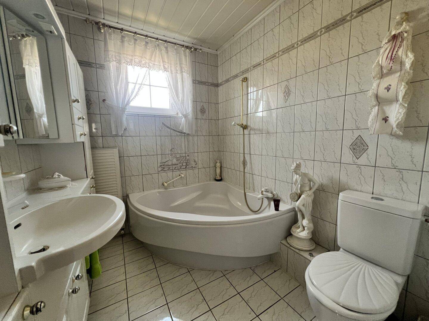 Gefliestes Badezimmer mit Eckbadewanne, Toilette, Waschbecken und Fenster.