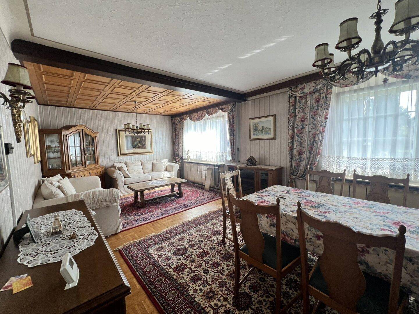Gemütlicher Wohn-Essbereich mit traditionellen Möbeln, Holzboden und großen Fenstern mit Vorhängen.