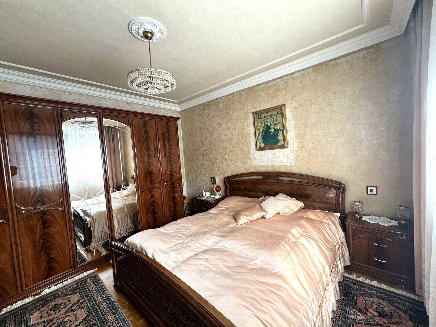 Schlafzimmer mit großem Holzbett, Spiegelschrank und gemusterter Tapete in warmen Farbtönen.
