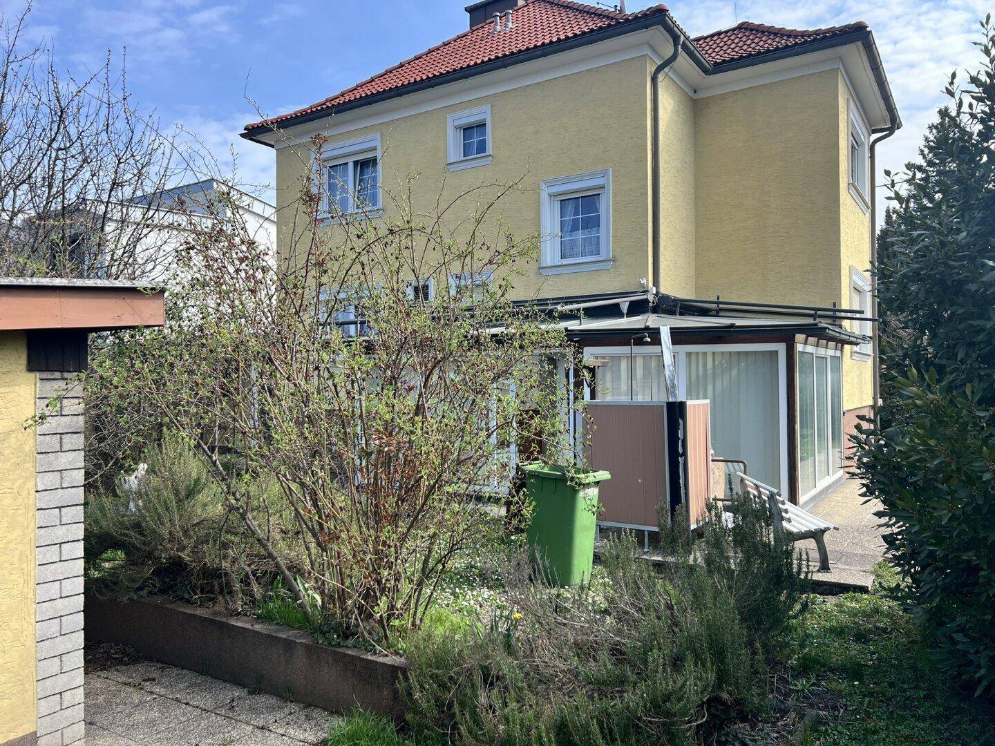 Gelbes Einfamilienhaus mit rotem Dach und einem Gartenbereich mit Sträuchern.