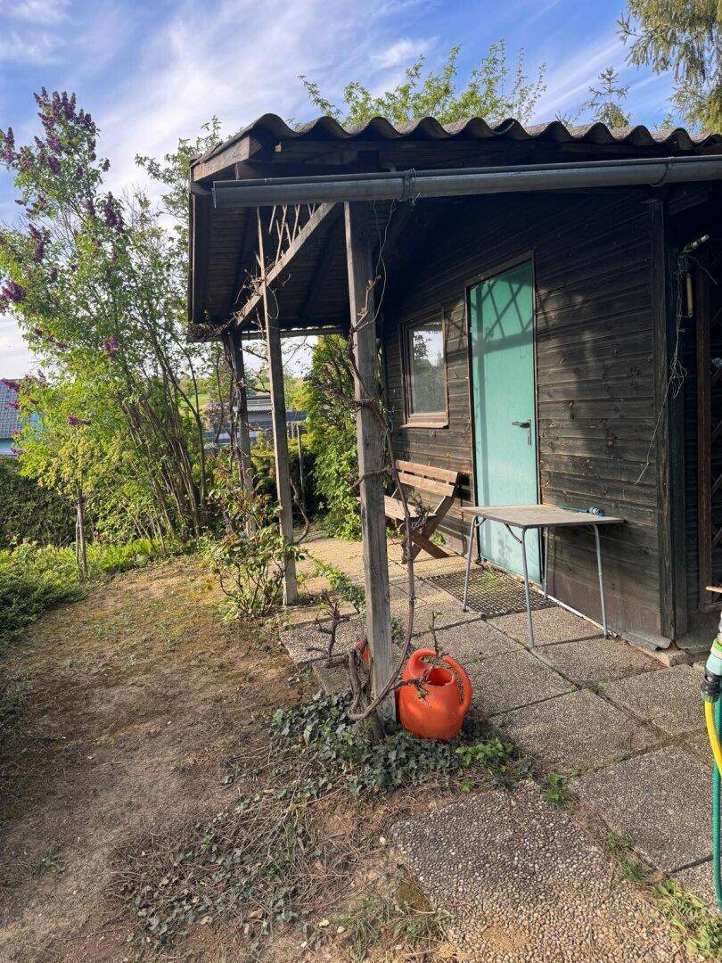 Rustikales Gartenhaus aus Holz mit kleiner überdachter Veranda und Sitzgelegenheit im Grünen.