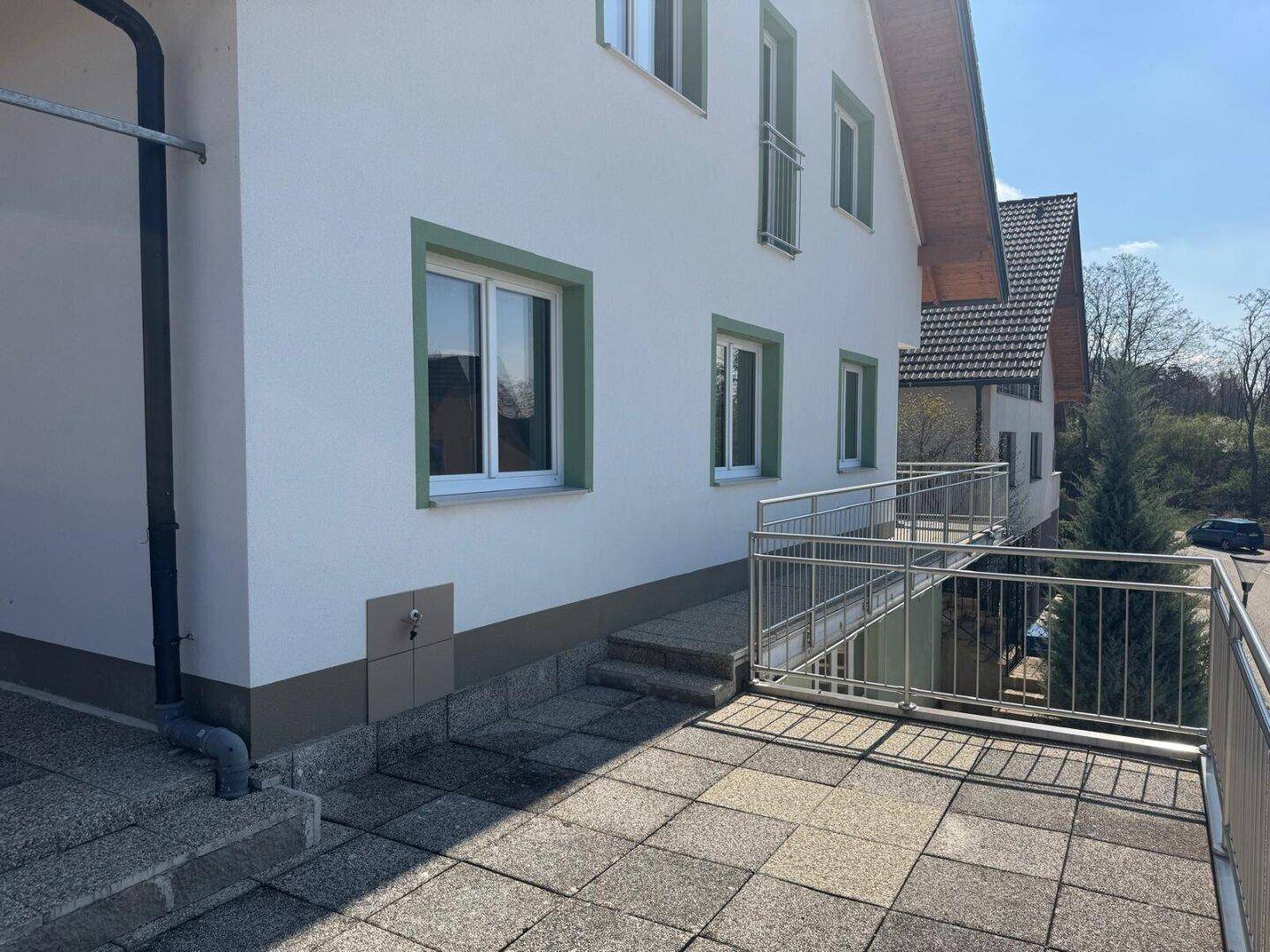 Moderne Hausfassade mit weißem Putz, grünen Fensterrahmen und einem gepflasterten Außenbereich mit Geländer.