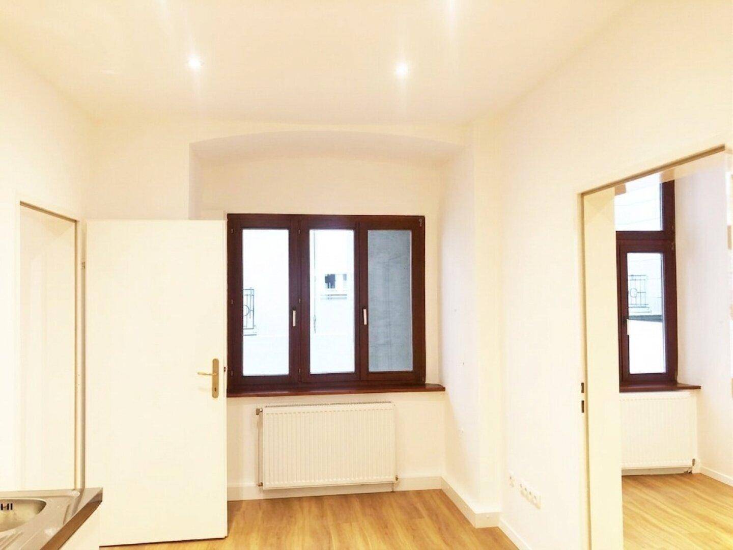 Heller Raum mit Holzboden, Fenster und Heizkörper, angrenzend an eine Küchenzeile.