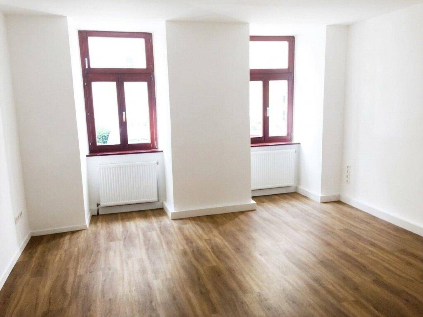 Leerer Raum mit zwei Fenstern, weißen Wänden und hellem Holzboden.