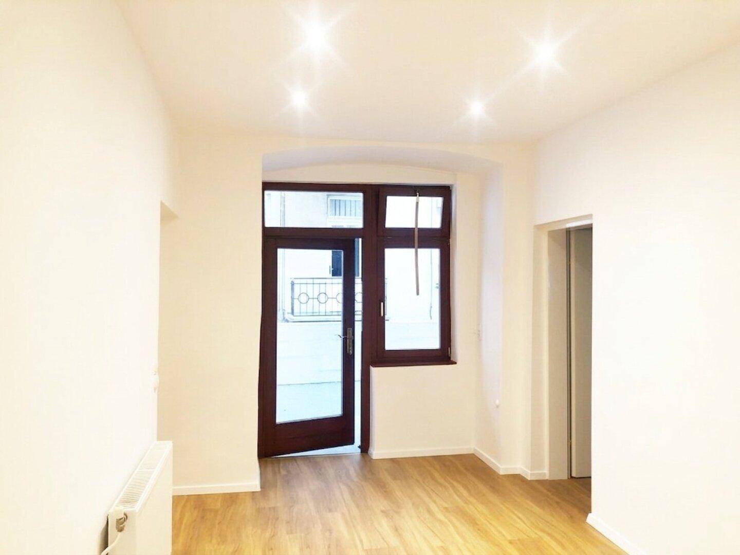 Heller Wohnraum mit Holzboden, weißen Wänden und einer Tür zu einem französischen Balkon.