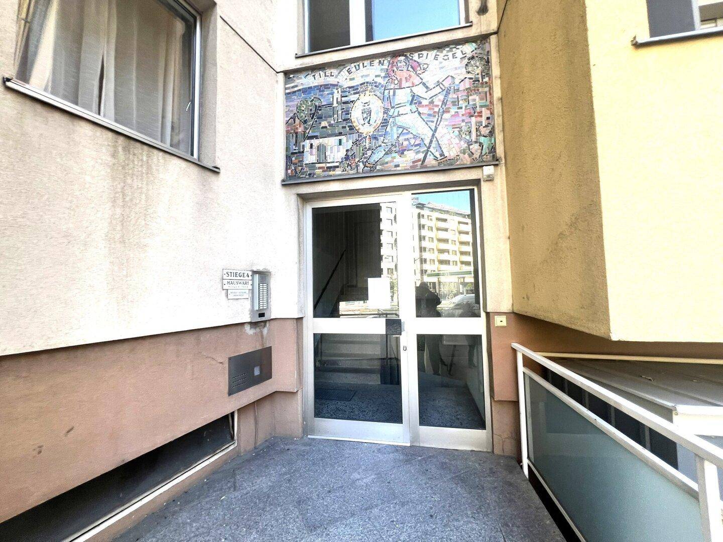 Gebäudeeingang mit Glastür, künstlerischem Mosaik und Zugang zur Straße.