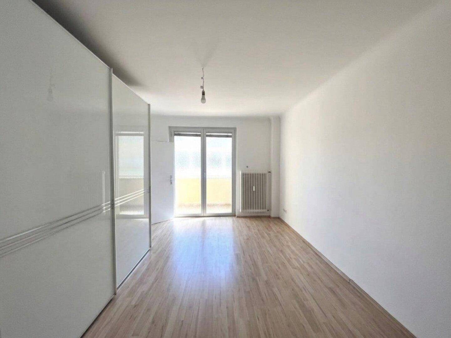 Lichtdurchflutetes Zimmer mit Laminatboden, weißem Schrank und Balkonzugang.