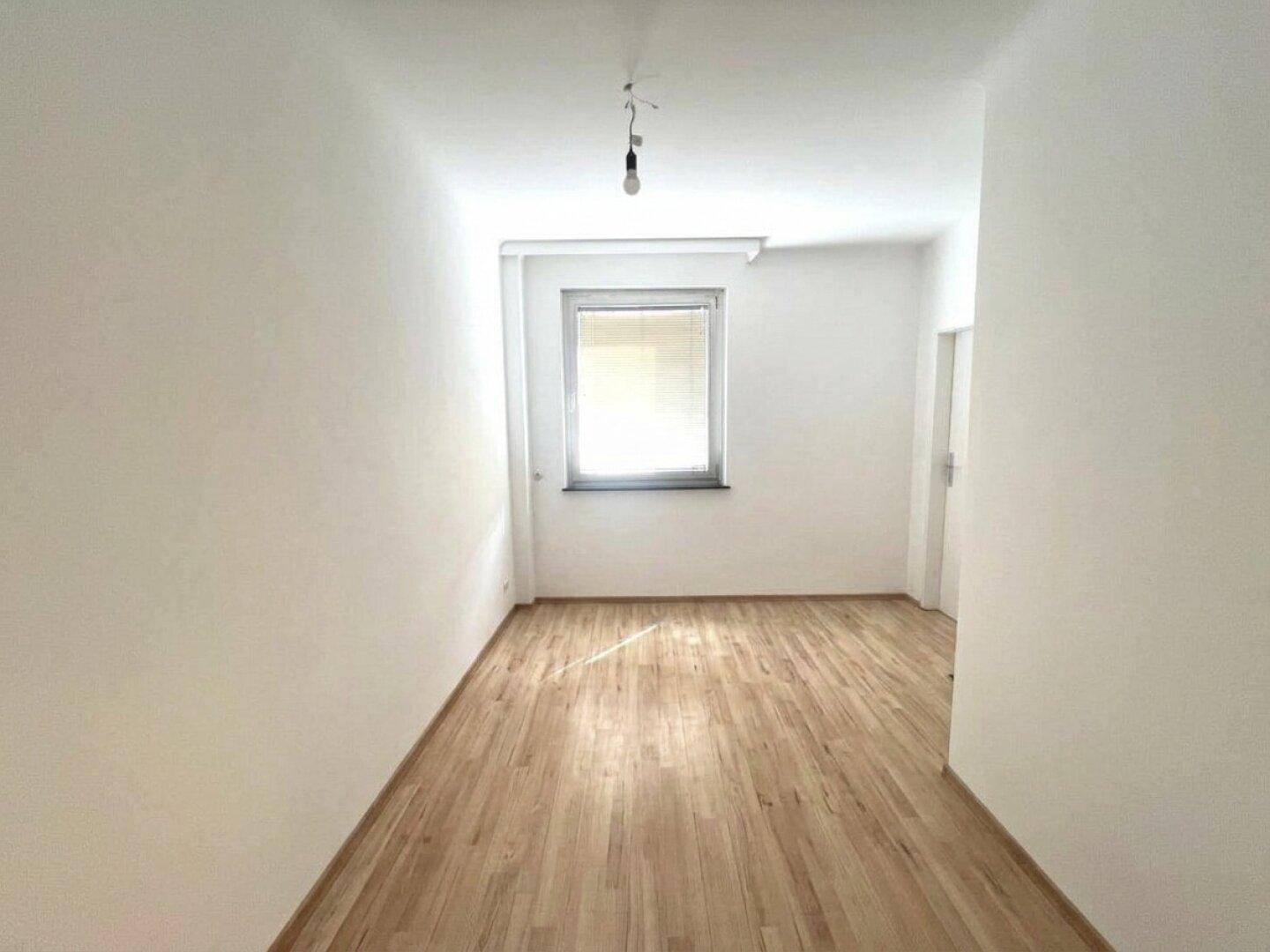 Helles Zimmer mit Laminatboden und einem großen Fenster, das viel Tageslicht hereinlässt.
