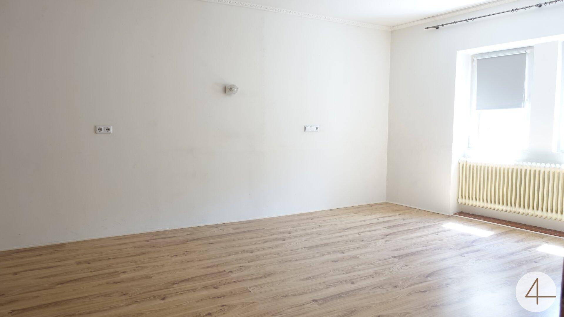 Heller, leerer Raum mit Laminatboden, weißen Wänden und einem Fenster mit Heizkörper.