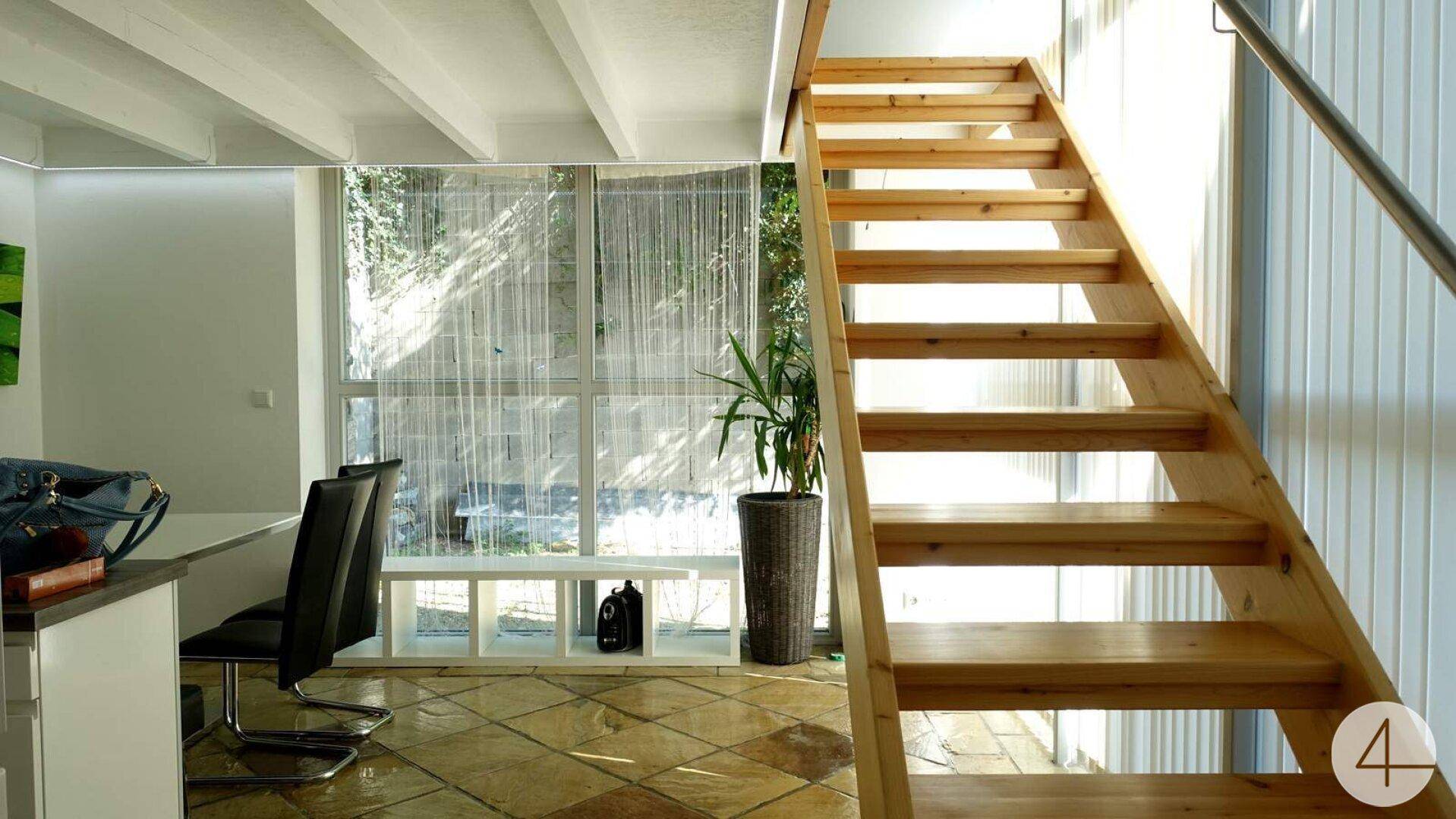 Helle Holztreppe mit Geländer in einem lichtdurchfluteten Bereich mit großen Fenstern.