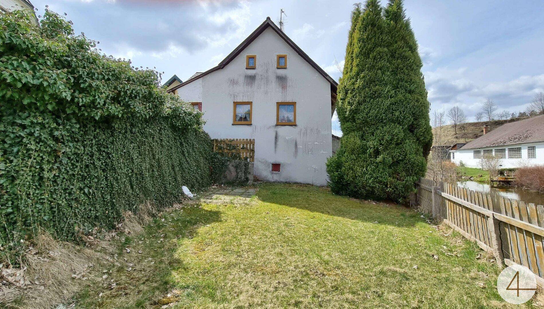 Hinterhof mit Rasenfläche, umgeben von einer Hecke und einem Holzzaun unter blauem Himmel.