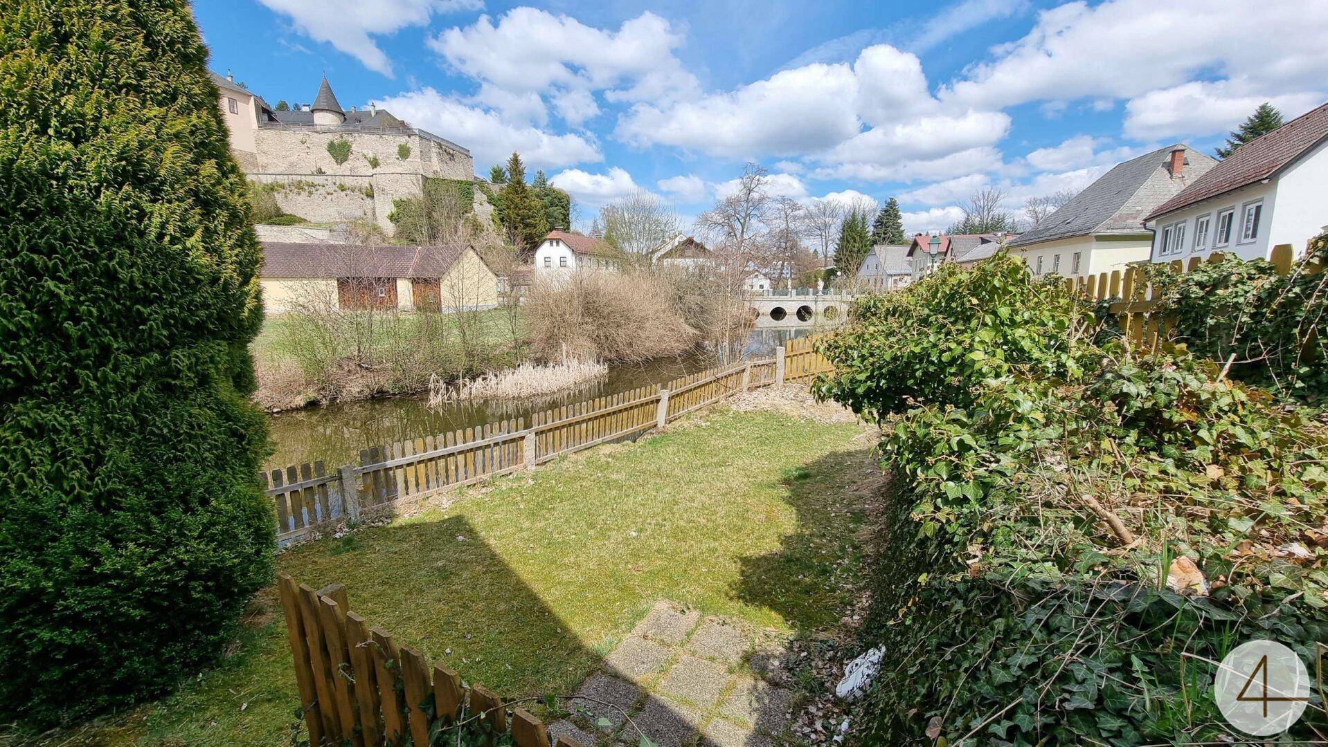 Gartenbereich mit Rasen, Holzzaun und Blick auf einen Fluss oder See sowie eine Brücke.