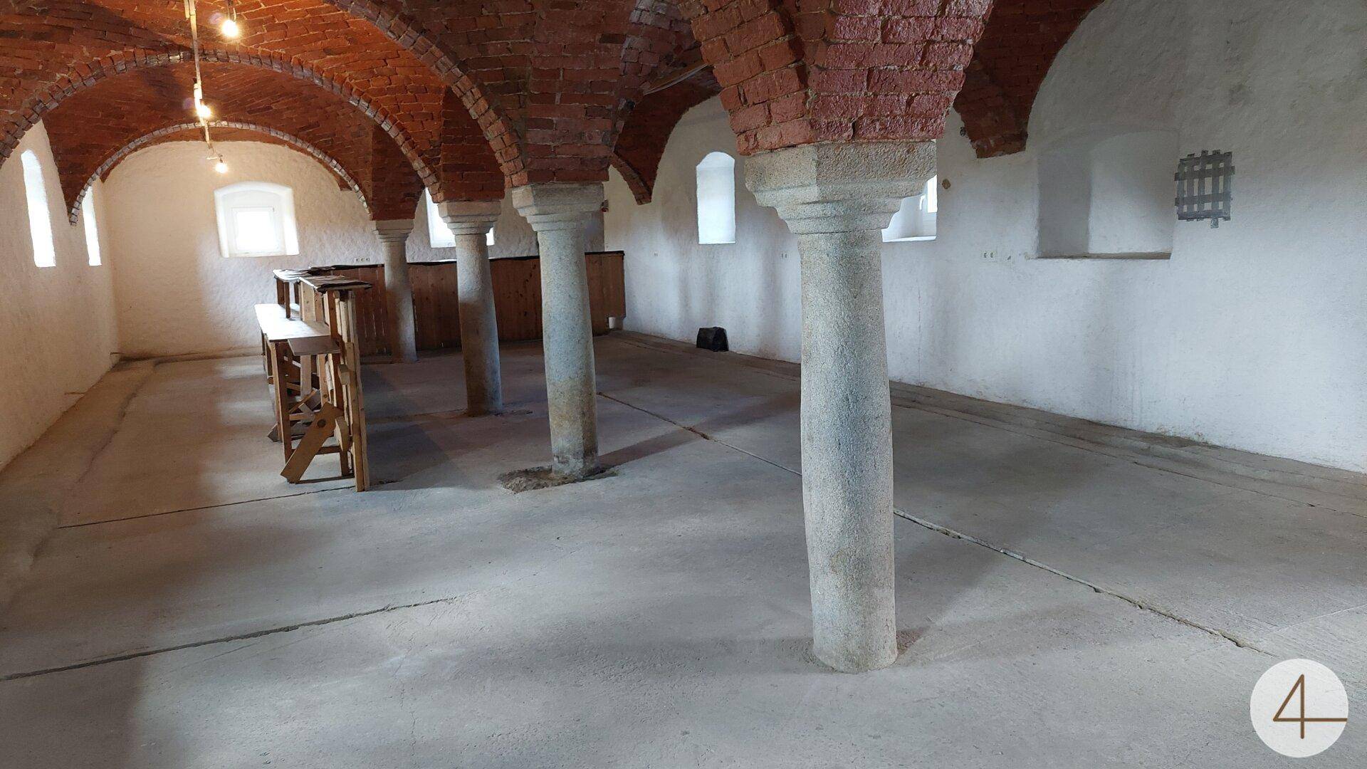 Historischer Gewölbekeller mit Ziegelbögen, Steinsäulen und kleinen Fenstern, hell beleuchtet.