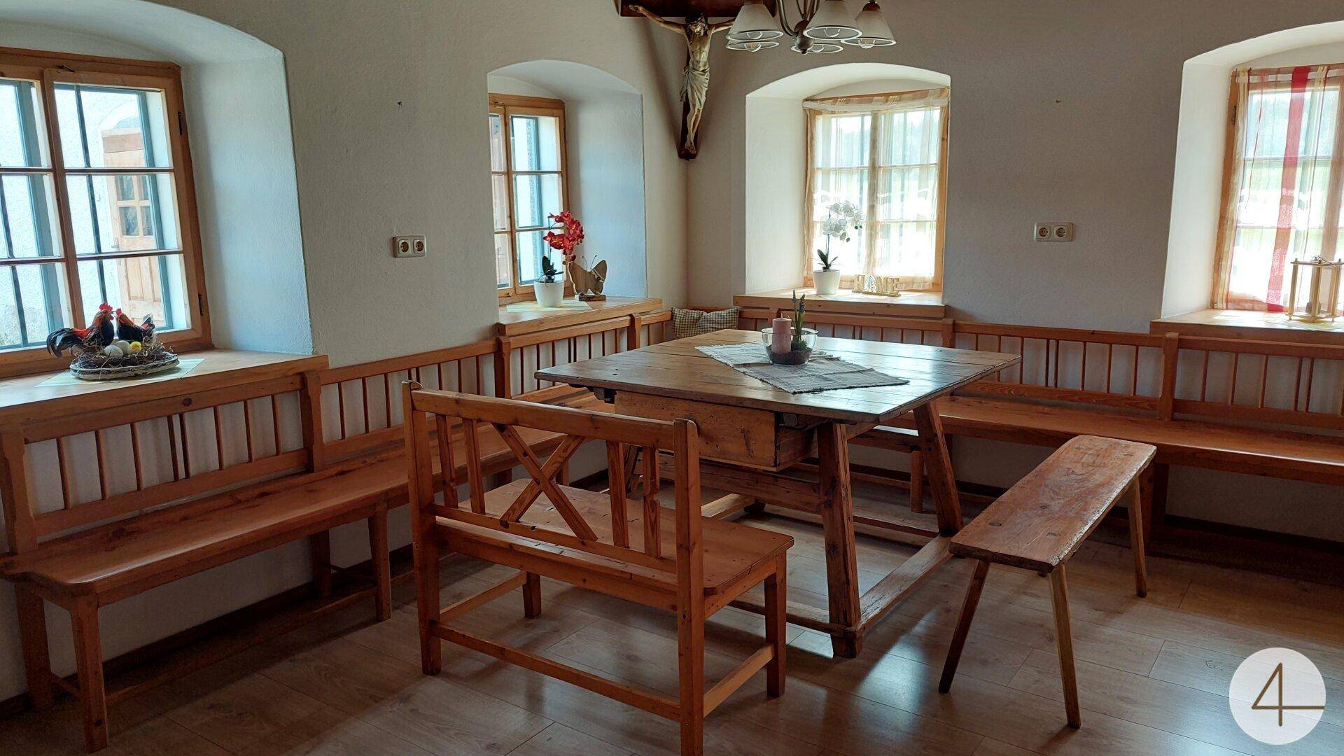 Traditioneller Essbereich mit Holztisch und Bänken, umgeben von Fenstern mit Vorhängen.