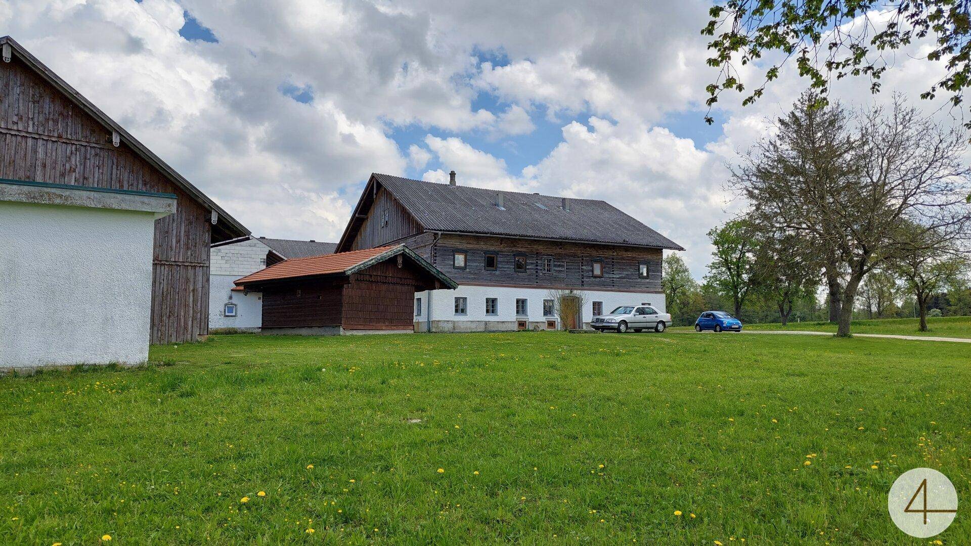 Weitläufige grüne Wiese mit traditionellen Gebäuden und Bäumen unter bewölktem Himmel.