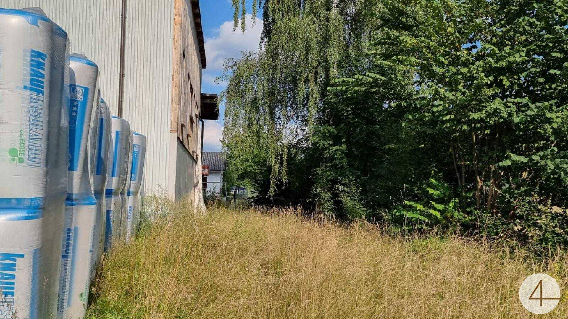 Hohes Gras wächst neben einem Gebäude, daneben gestapelte Isolationsmaterialien und Bäume.