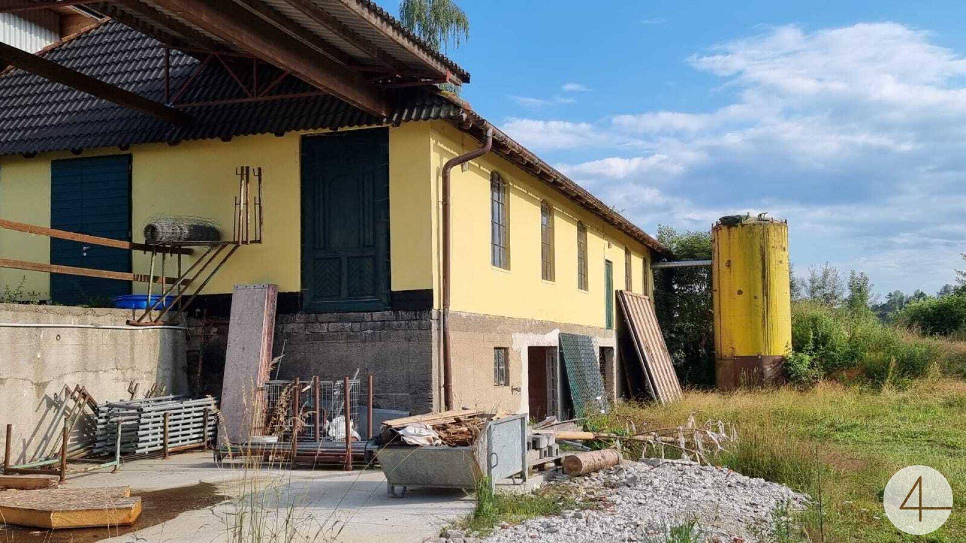 Altes gelbes Gebäude mit dunklen Fensterläden und einem gelben Silo im ländlichen Umfeld.