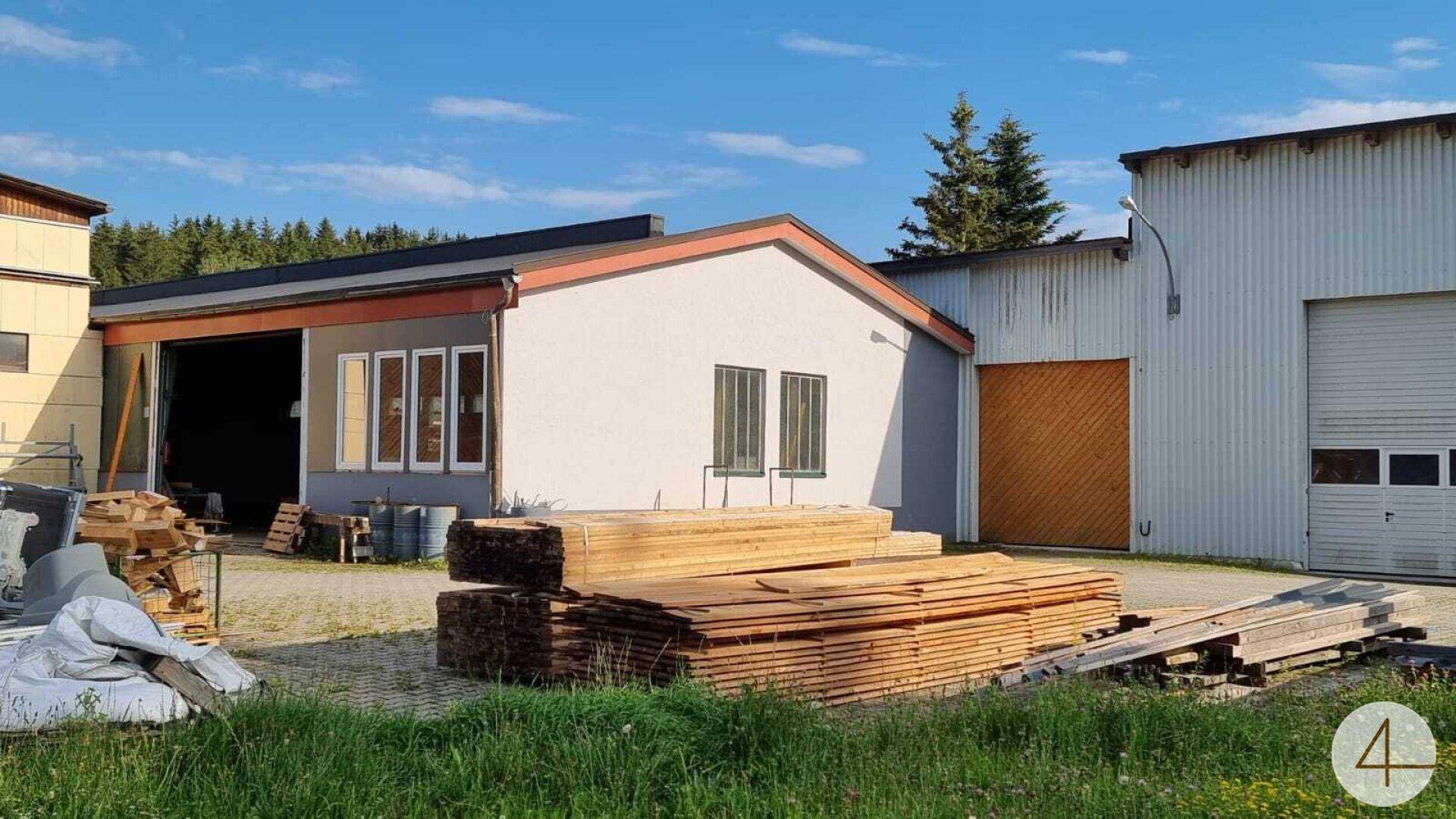 Mehrere Industriegebäude mit gestapeltem Holz auf einem Schotterplatz unter blauem Himmel.
