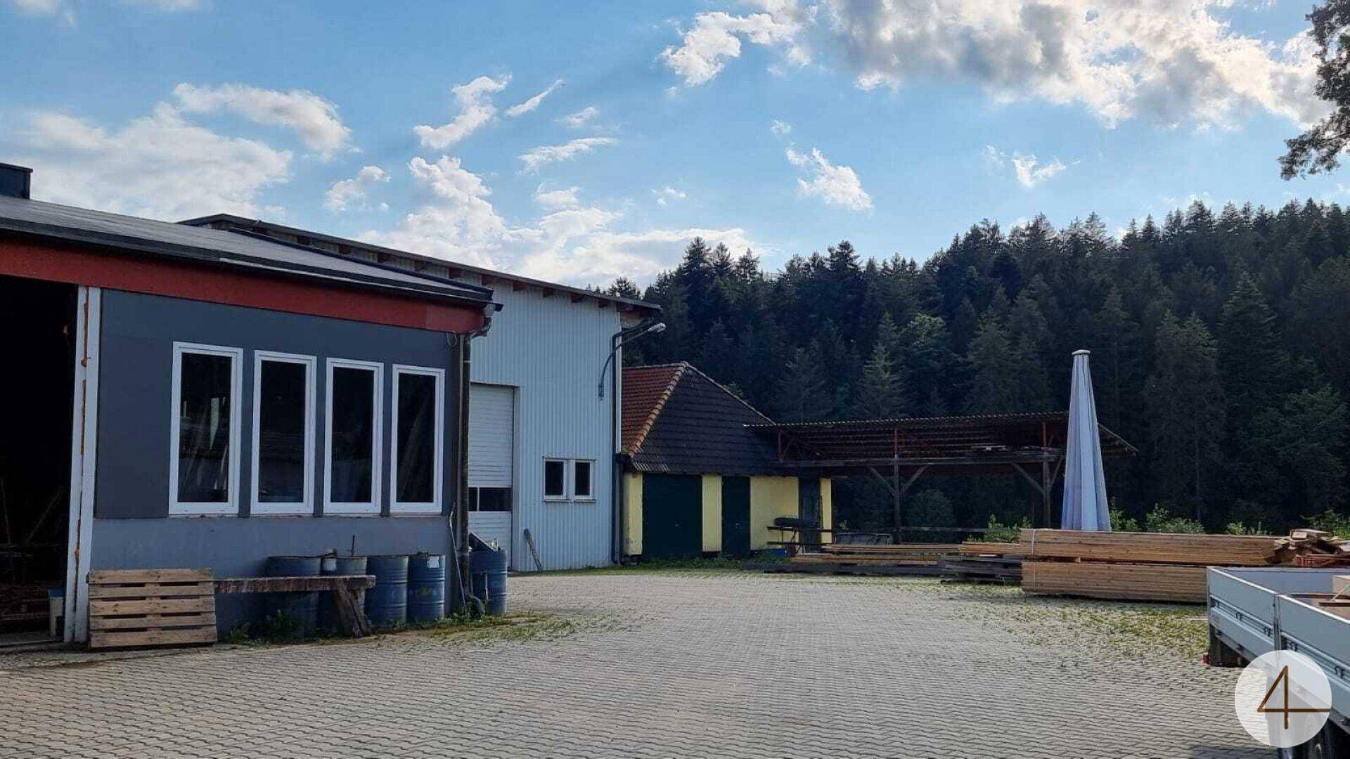 Großer gepflasterter Hofbereich zwischen verschiedenen Betriebsgebäuden mit Wald im Hintergrund.