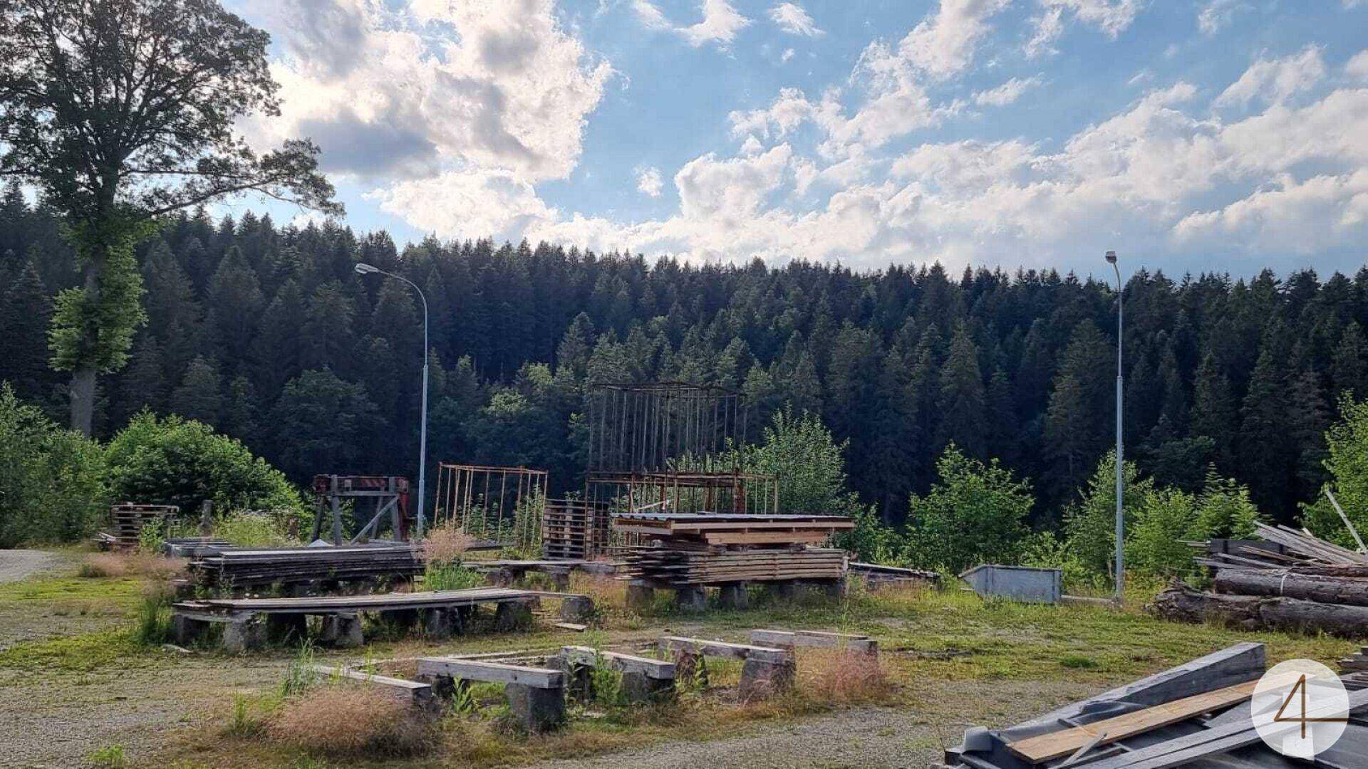 Offener Lagerplatz mit Holzstapeln und Metallkonstruktionen vor einem dichten Wald.