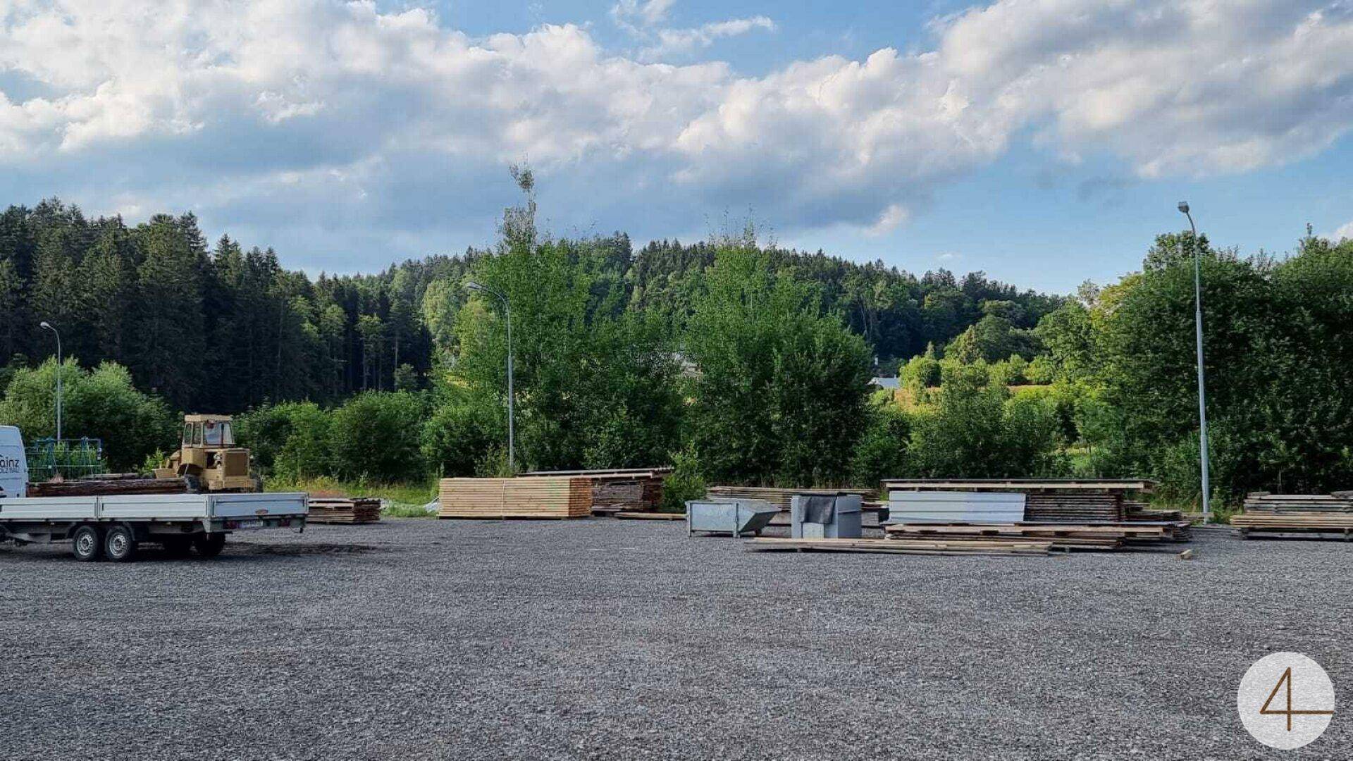 Großer Schotterplatz mit gestapeltem Holz und Baumaterialien, umgeben von Bäumen und Wald.