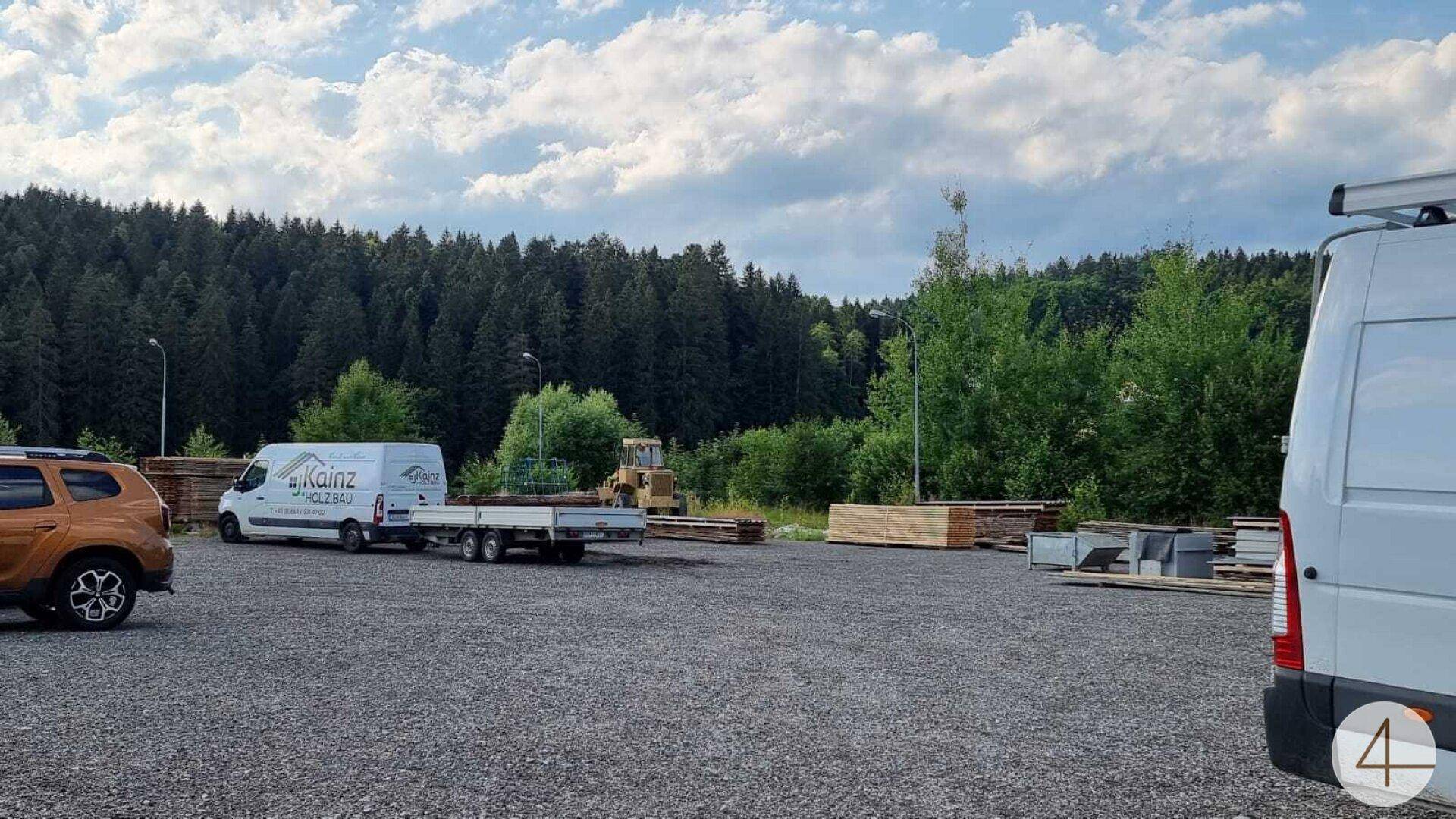 Großer Schotterparkplatz mit Lieferwagen und Anhänger, umgeben von Wald und grüner Landschaft.