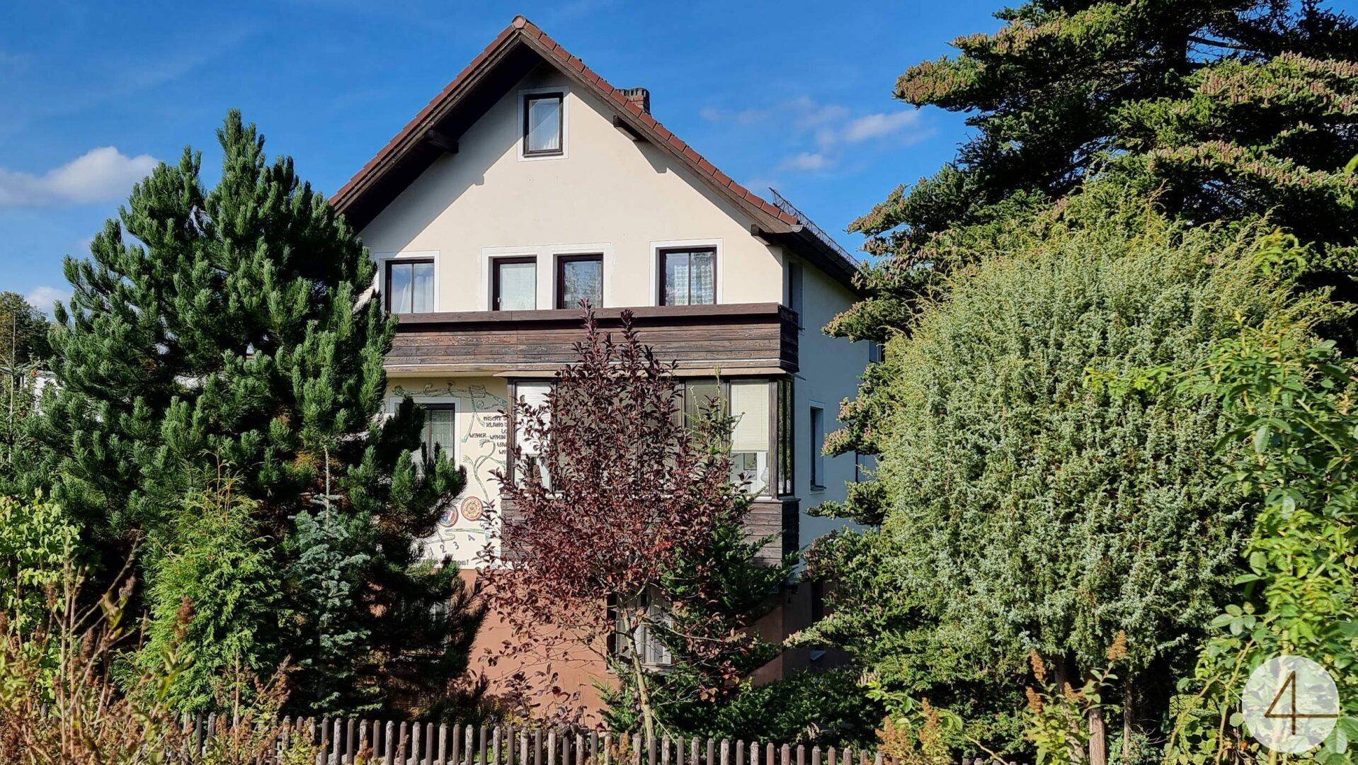 Ansicht der Hausfassade mit Satteldach, umgeben von üppiger Vegetation und einem Holzzaun.