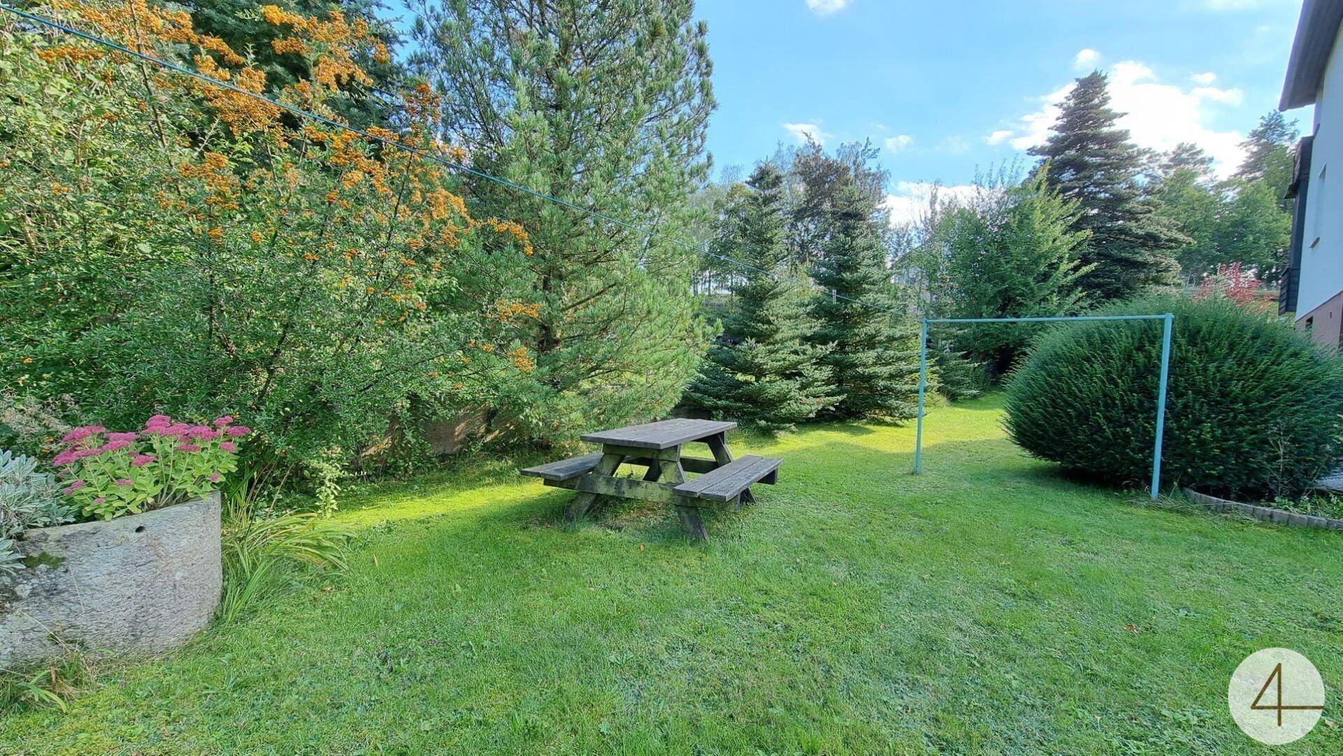 Gemütlicher Gartenbereich mit Holztisch und Bänken, umgeben von Grünflächen und Bäumen.