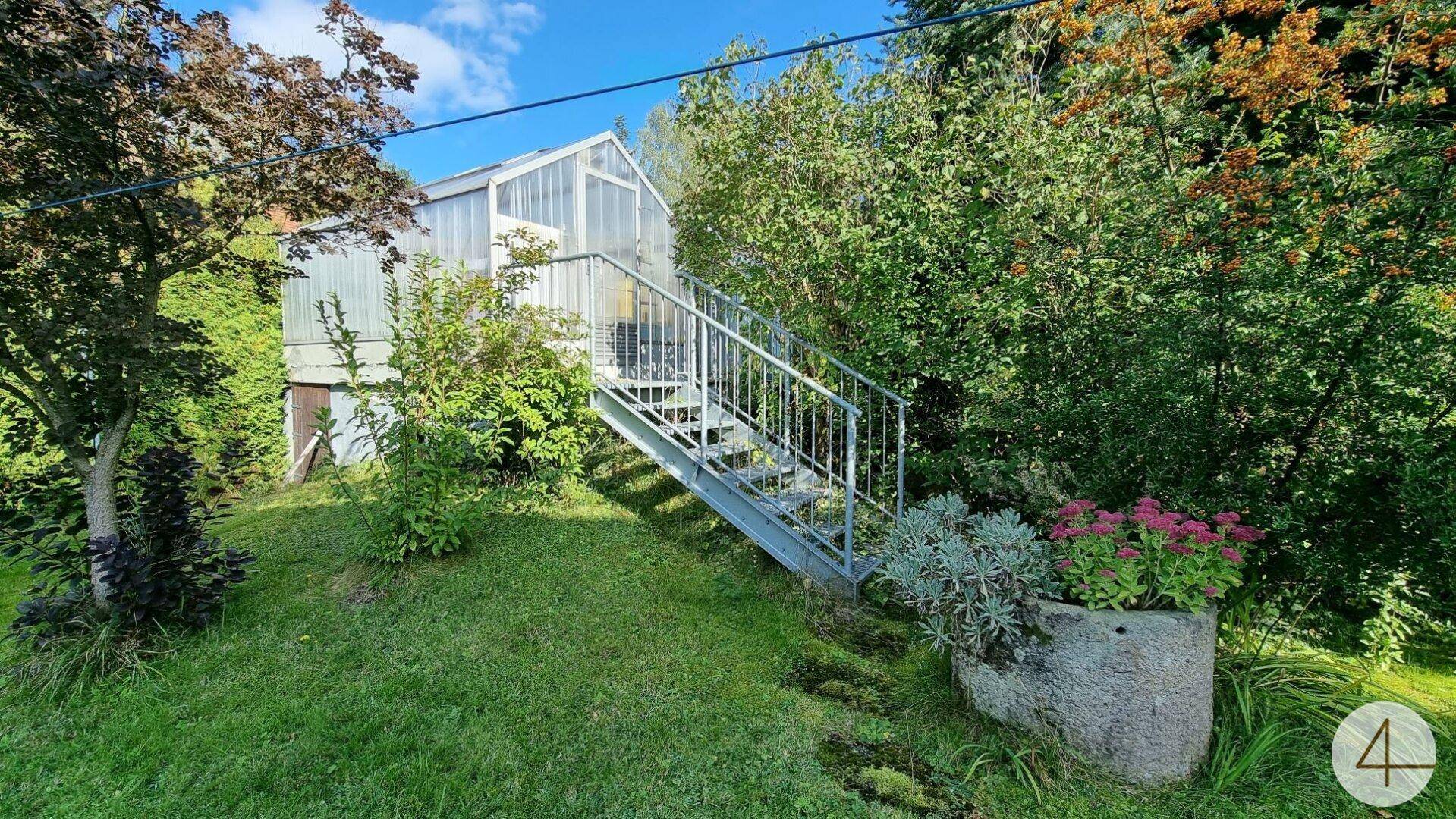 Garten mit einem Gewächshaus und einer Metalltreppe, umgeben von Pflanzen und Sträuchern.