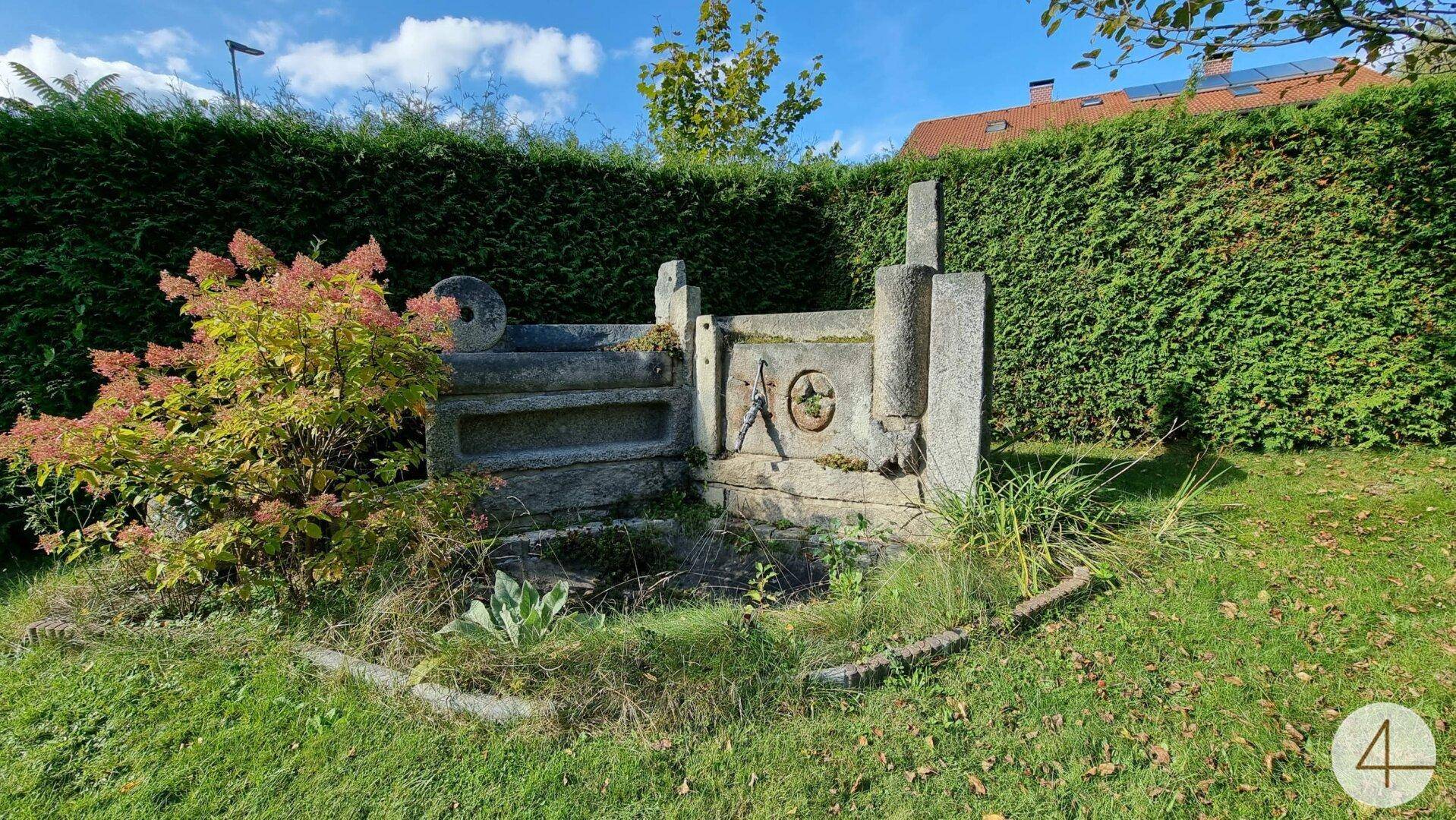 Gartenbereich mit einem alten Steinbrunnen oder einer dekorativen Steinstruktur und Hecke.