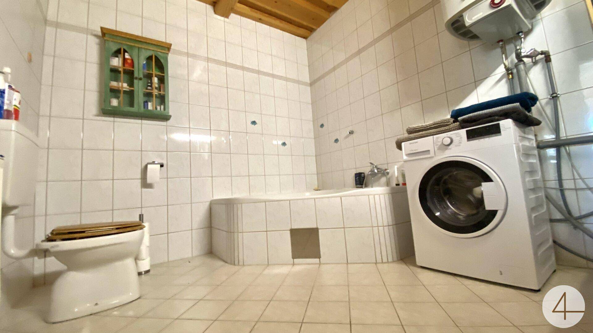 Gefliestes Badezimmer mit Badewanne, Toilette und Waschmaschine unter einer Holzdecke.