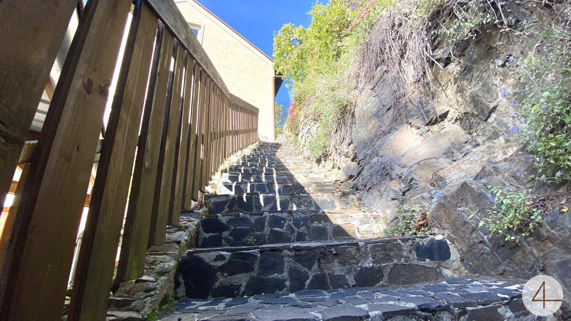 Steiler Steinweg mit Holzgeländer, der zu einem Gebäude hinaufführt, umgeben von Felsen und Vegetation.