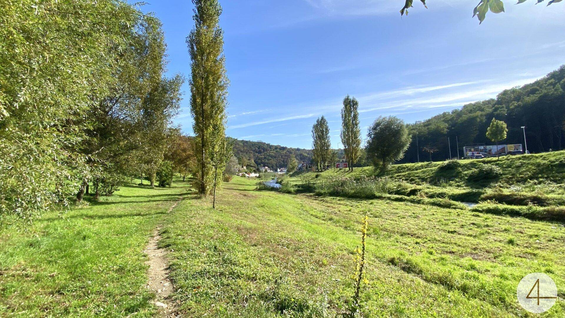 Weitläufige grüne Landschaft mit Bäumen, einem kleinen Bach und bewaldeten Hügeln unter blauem Himmel.