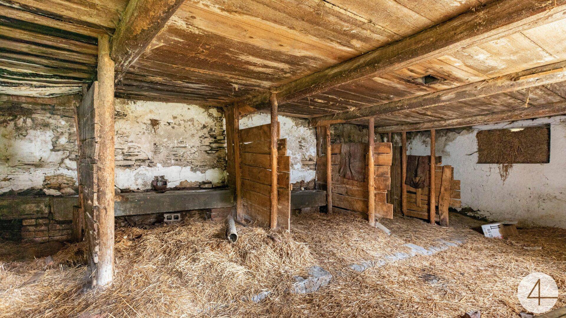 Rustikaler Innenraum eines alten Stalls mit Holzbalken, Stroh auf dem Boden und verwitterten Wänden.