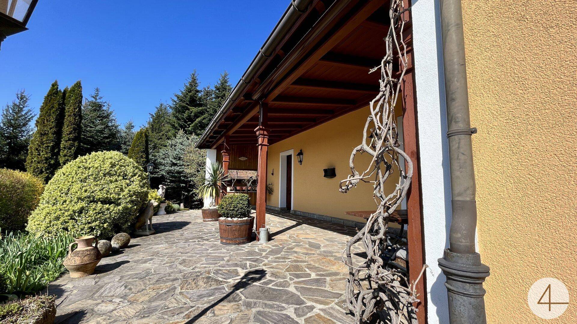 Überdachte Terrasse mit rustikalen Holzsäulen und Steinplattenboden, umgeben von viel Grün.