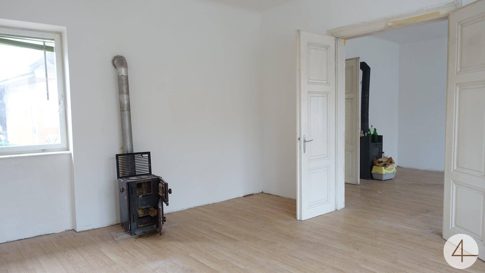 Helles Wohnzimmer mit Holzboden, weiß gestrichenen Wänden und einem traditionellen Holzofen als Heizquelle.