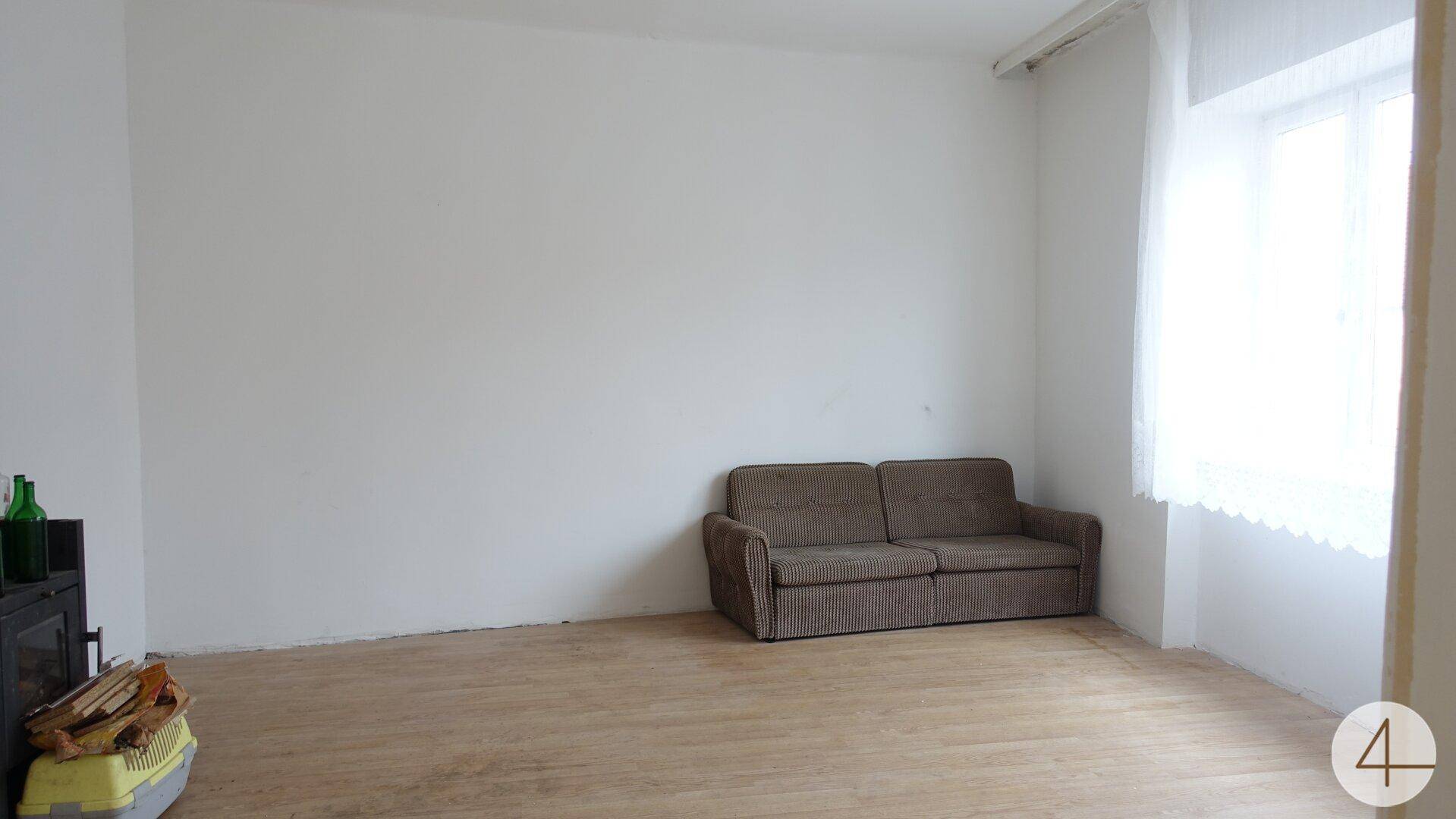 Geräumiges Wohnzimmer mit hellem Holzboden, weiß gestrichenen Wänden und einem bequemen braunen Sofa.