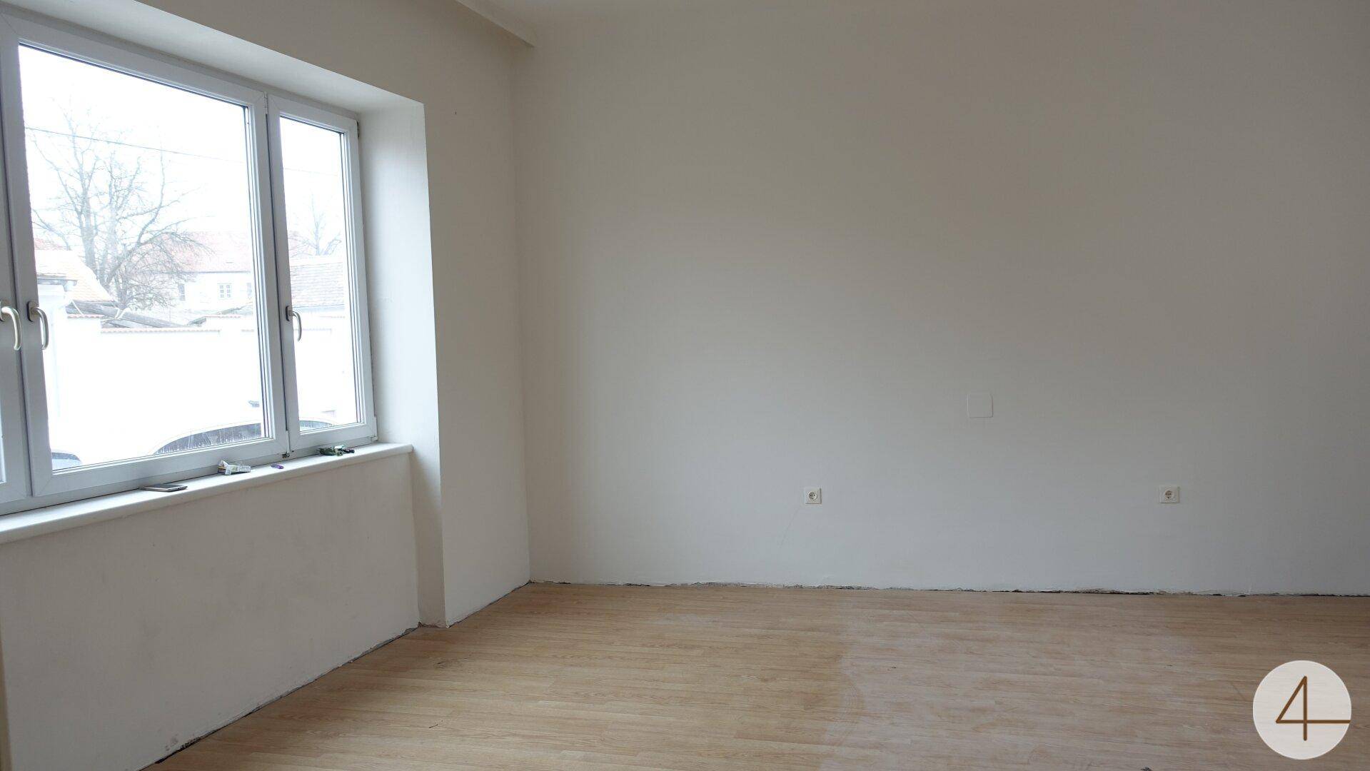 Heller Raum mit großen Fenstern, die viel Tageslicht hereinlassen, und einem einfachen Holzboden.