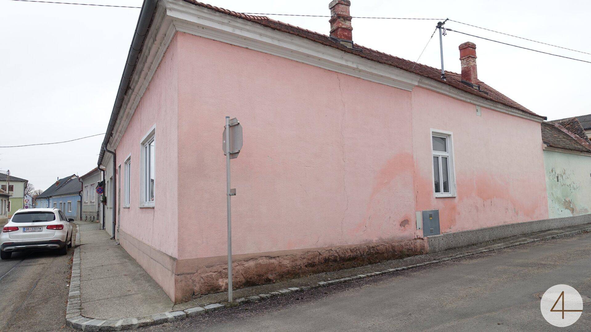 Seitenansicht des rosafarbenen Hauses an einer Straßenecke mit einem geparkten Auto und weiteren Gebäuden im Hintergrund.
