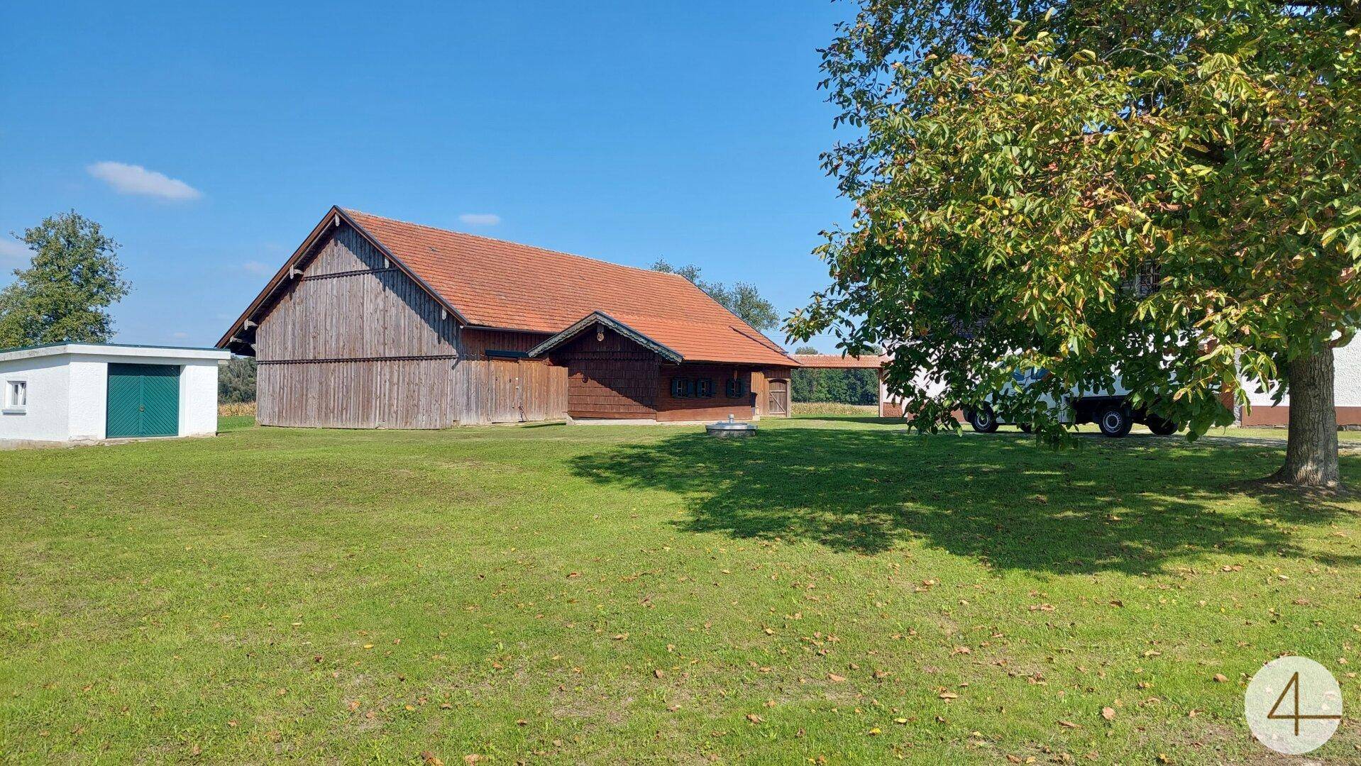 Weitläufige grüne Wiese mit einem Holzgebäude und einem kleinen Schuppen unter blauem Himmel.