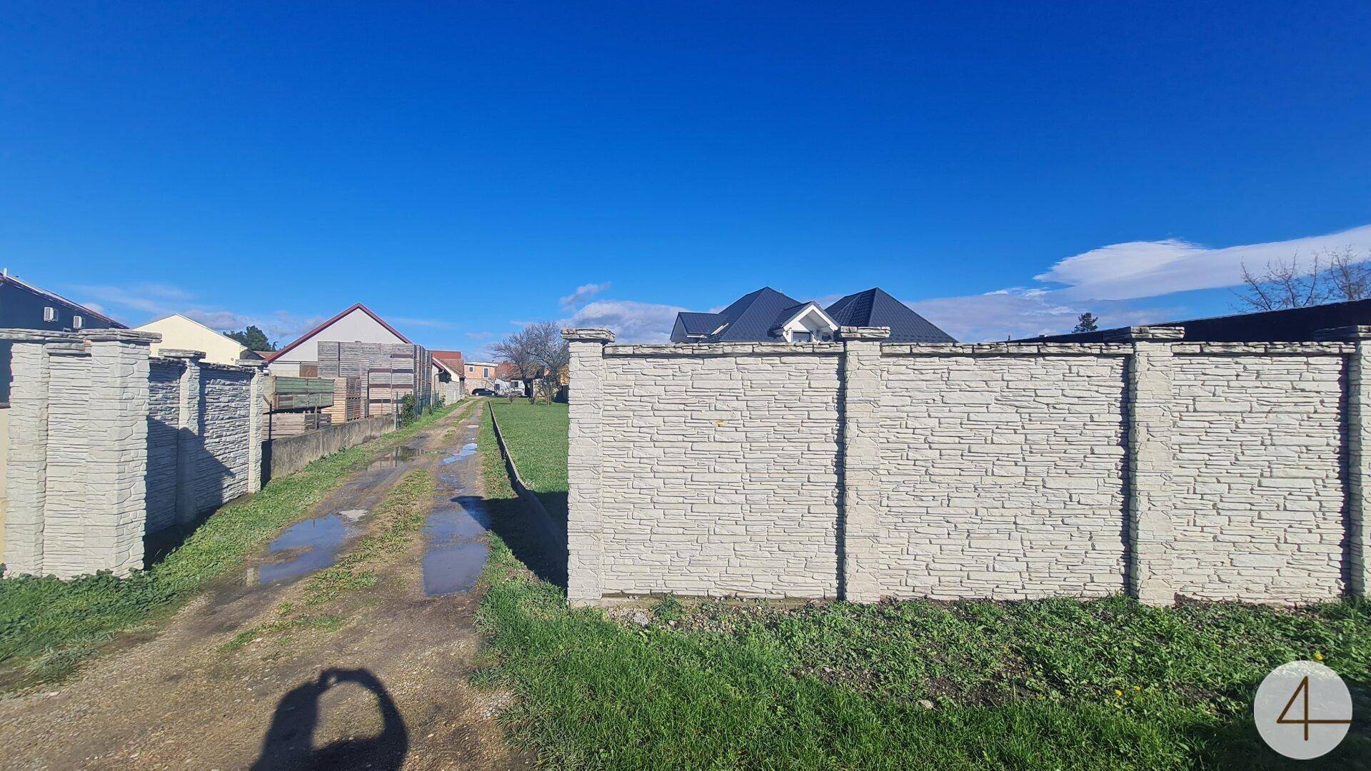 Unbefestigter Weg führt an einer dekorativen Steinmauer vorbei, mit weiteren Häusern und blauem Himmel im Hintergrund.