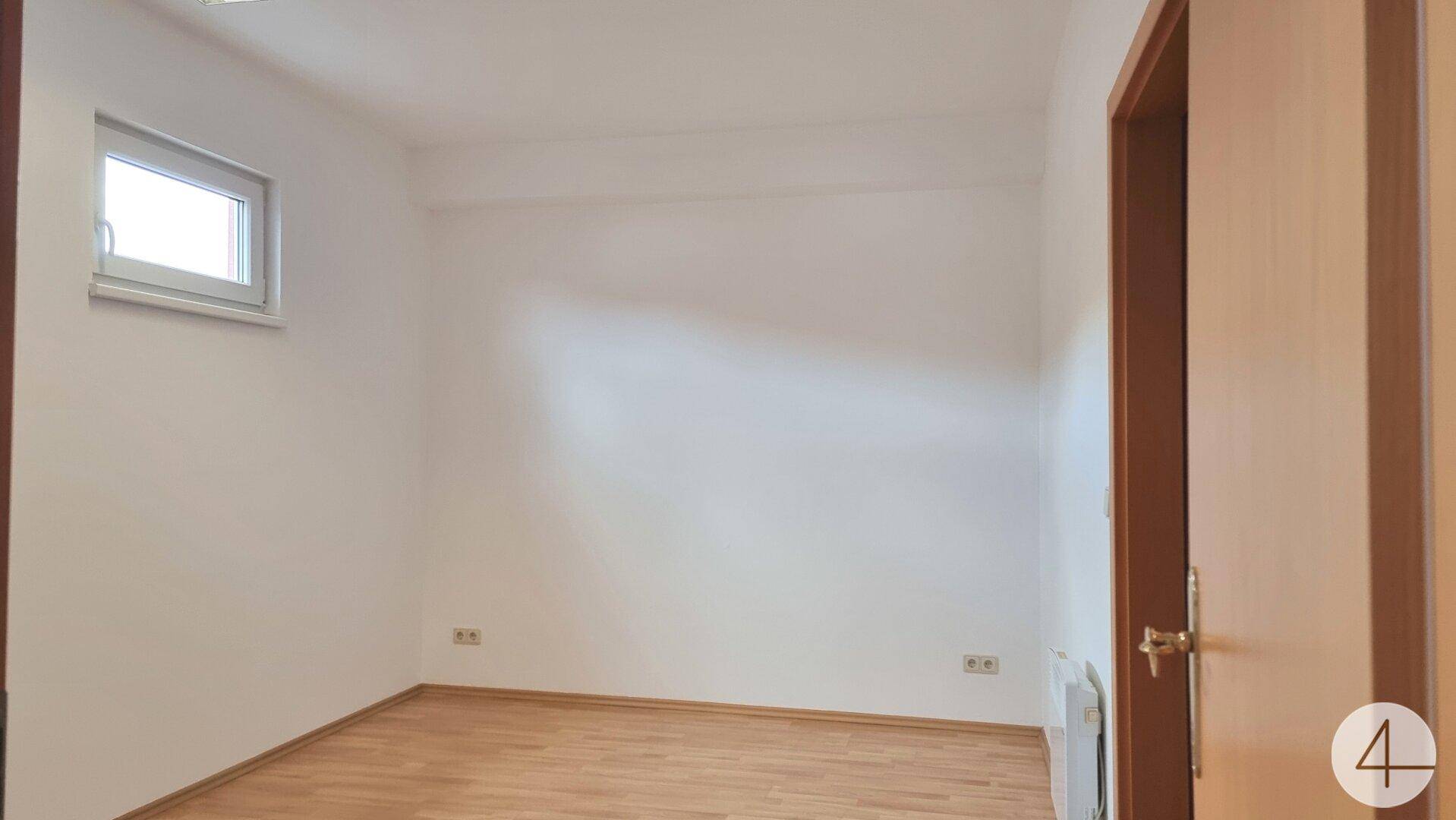 Leerer Raum mit Laminatboden und einem Fenster, das Tageslicht hereinlässt, ideal für ein Schlafzimmer oder Büro.