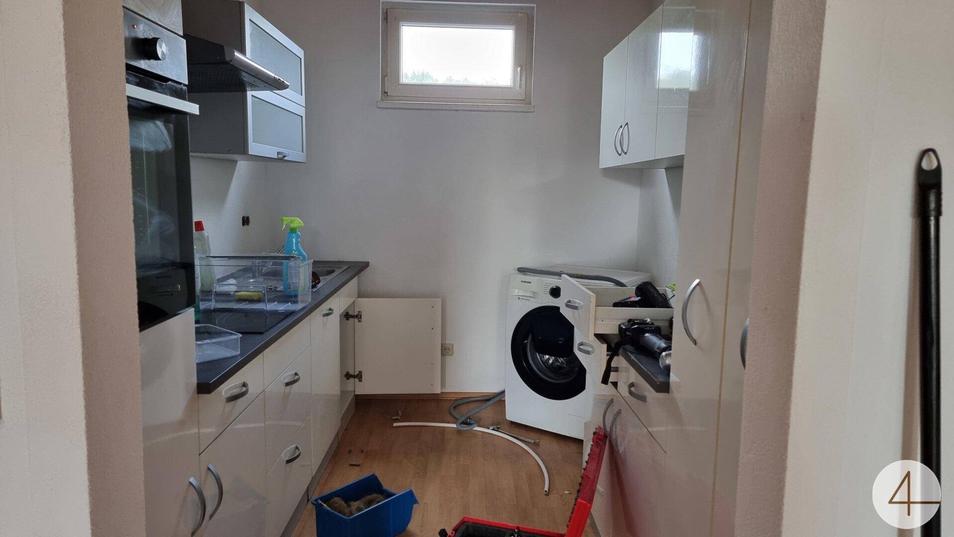 Küche mit weißen Schränken, Einbaugeräten und einer Waschmaschine, die auf den Einbau wartet.