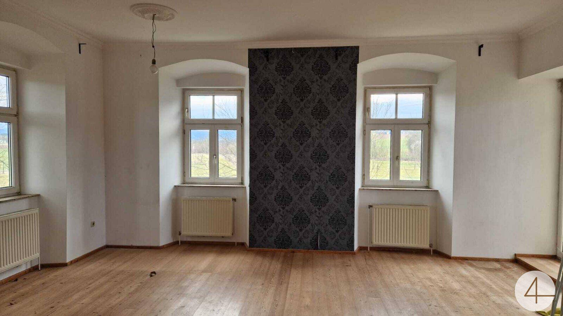 Geräumiger Raum mit Holzdielenboden, hohen Fenstern und einer Akzentwand mit Tapete.