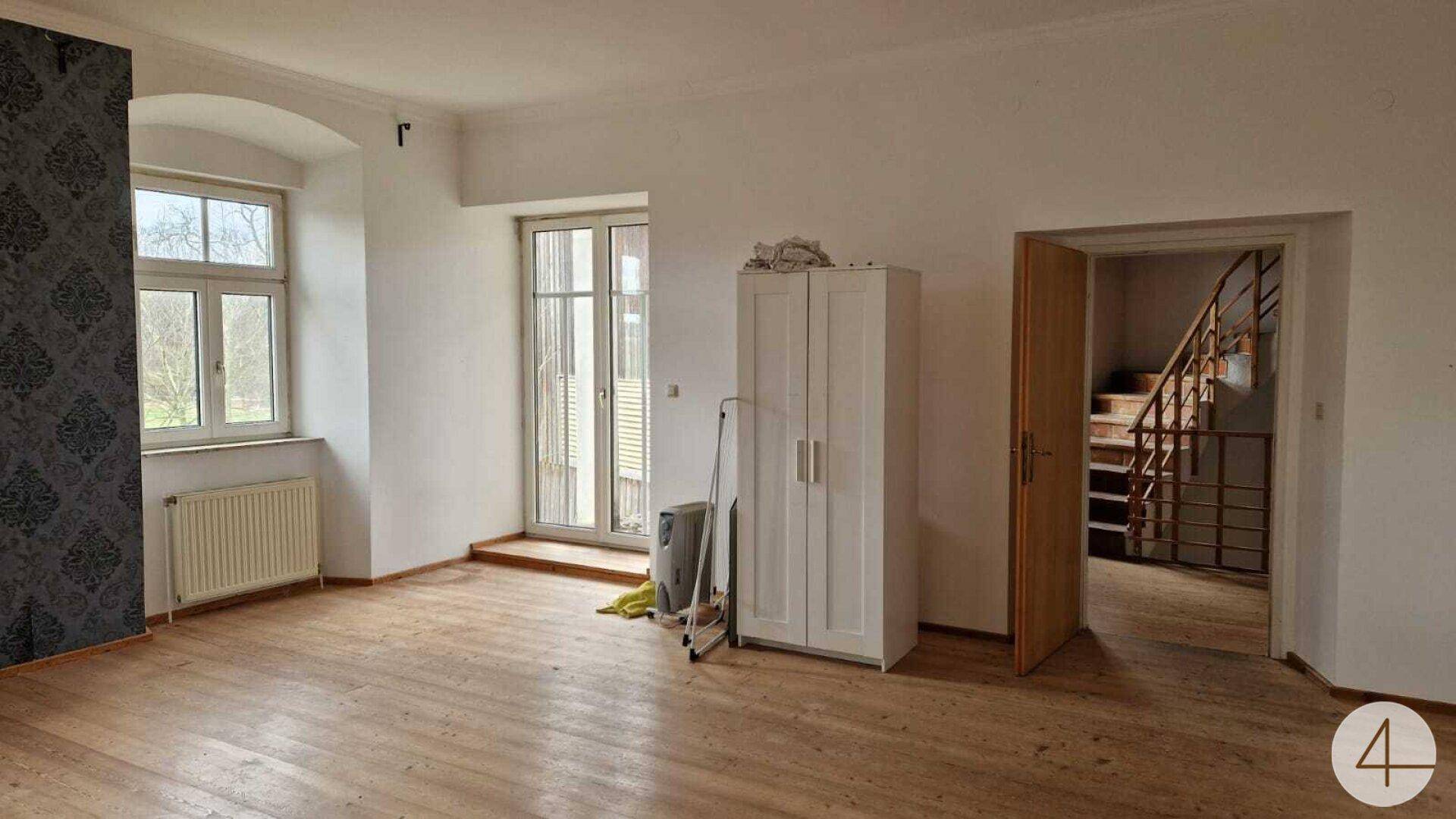 Geräumiger Raum mit Holzdielenboden, hohen Fenstern und Zugang zu einem weiteren Bereich.
