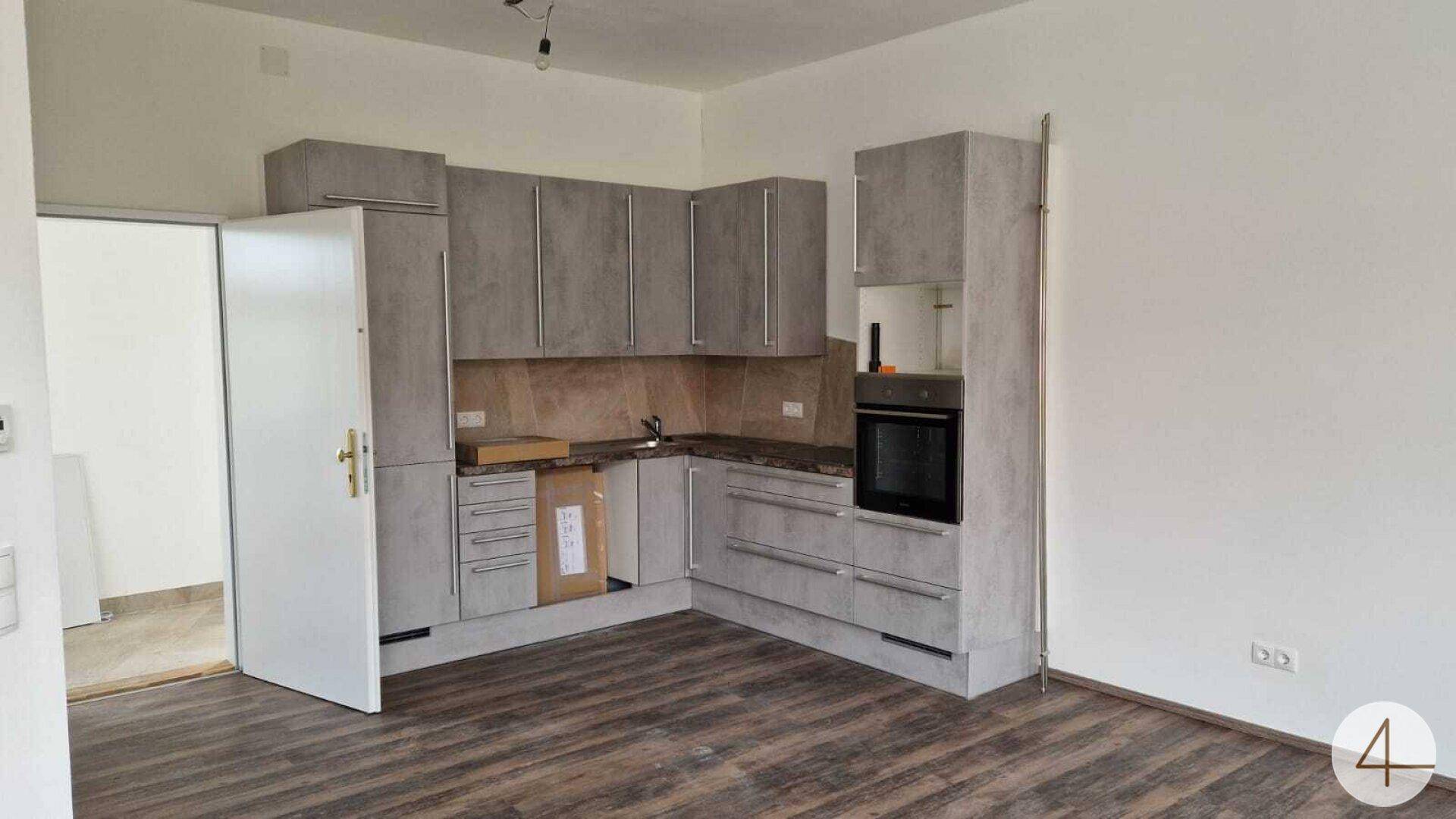 Moderne Einbauküche in Grau mit integriertem Backofen und dunklem Laminatboden in einem hellen Raum.