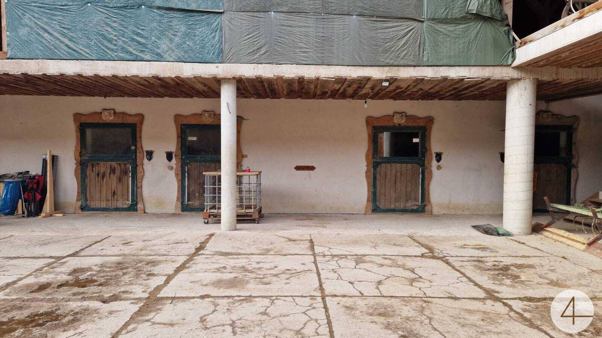 Außenansicht eines Stallgebäudes mit mehreren Holztüren zu den Pferdeboxen und einem gepflasterten Vorplatz.