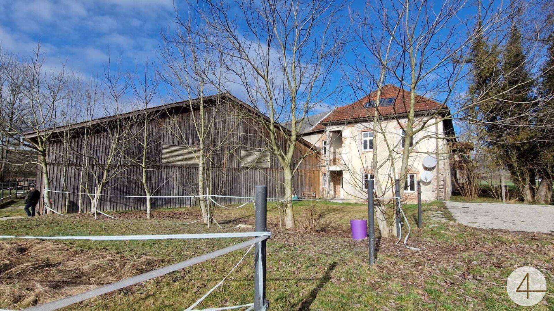 Zwei Gebäude, eine Holzscheune und ein traditionelles Haus, umgeben von Feldern und Bäumen.